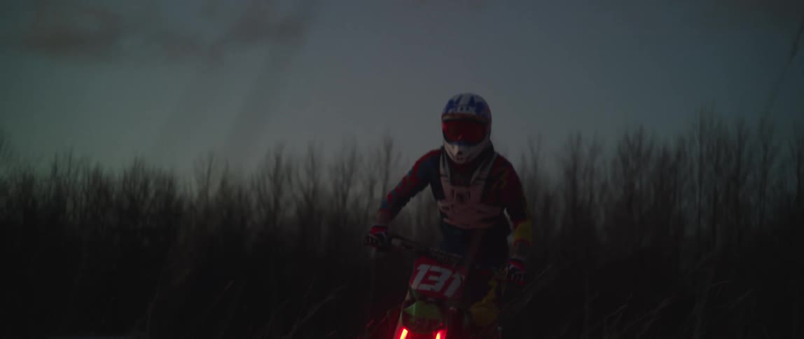 A cyclist wearing a helmet is riding a motorcycle with red lights through a field and does a wheelie