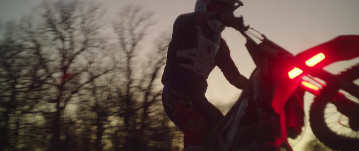 A motorcyclist stands and pops a wheelie as he rides through a field of tall grass at sunset with glowing red lights on his bike