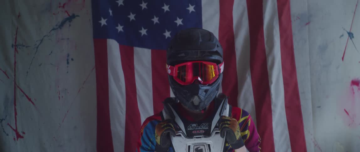 A person standing in a room wearing a racing helmet goggles and vest with an american flag in the background