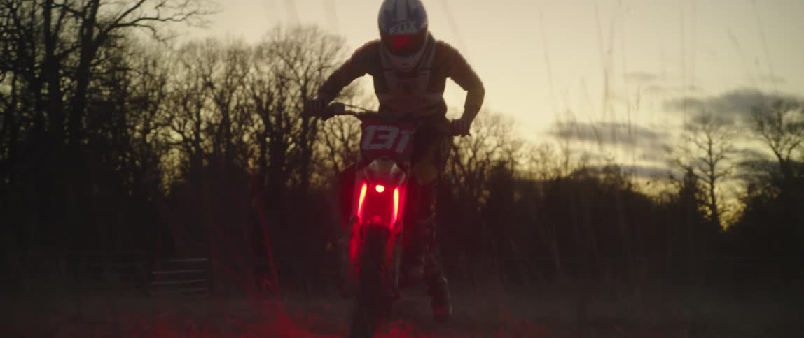 Person wearing helmet performs trick riding on a glowing red motocross bike in field near farmhouse