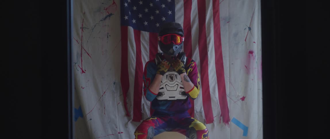 A racer sits posed in front of an american flag with racing helmet goggles and vest on