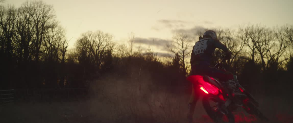 Motorcycle rider spins motorcycle with glowing red lights in small circles in field enclosed by fence and leafless trees