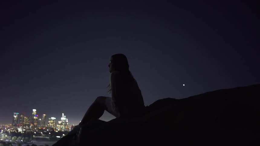 A woman is sitting on a hilltop taking in a view of a city at night
