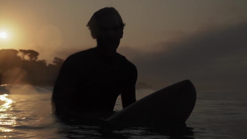 A man is floating in the water on a surfboard at dawn