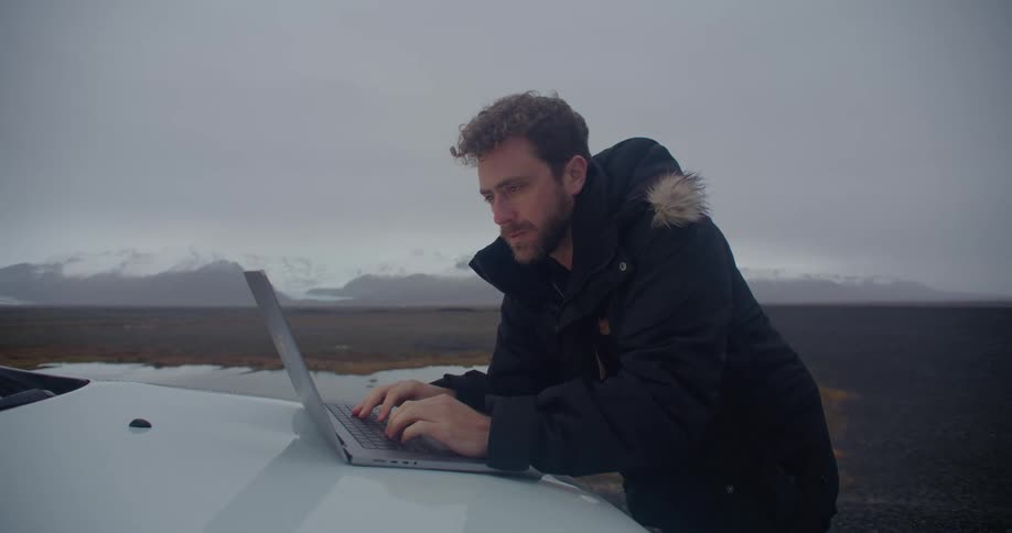 A man wearing a winter coat is typing on a laptop computer sitting on the front of an automobile and there are snow covered mountains in the distance