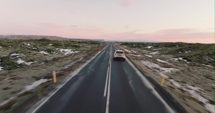 A white automobile is traveling on a road with light snow on the roadside and mountains on the horizon