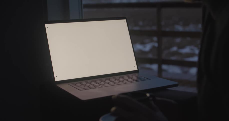 A laptop computer sits on a desk in a dark room near a window