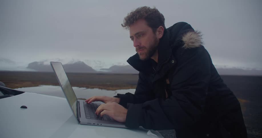 Man leans over hood of car parked near shore typing on laptop