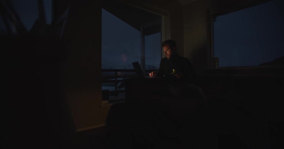 Man with beard black sweater sitting at table uses laptop in dark room