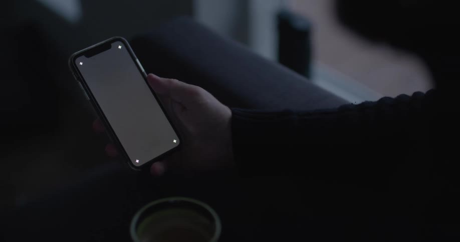 Man with black sweatshirt holds cup and uses smartphone in dark room