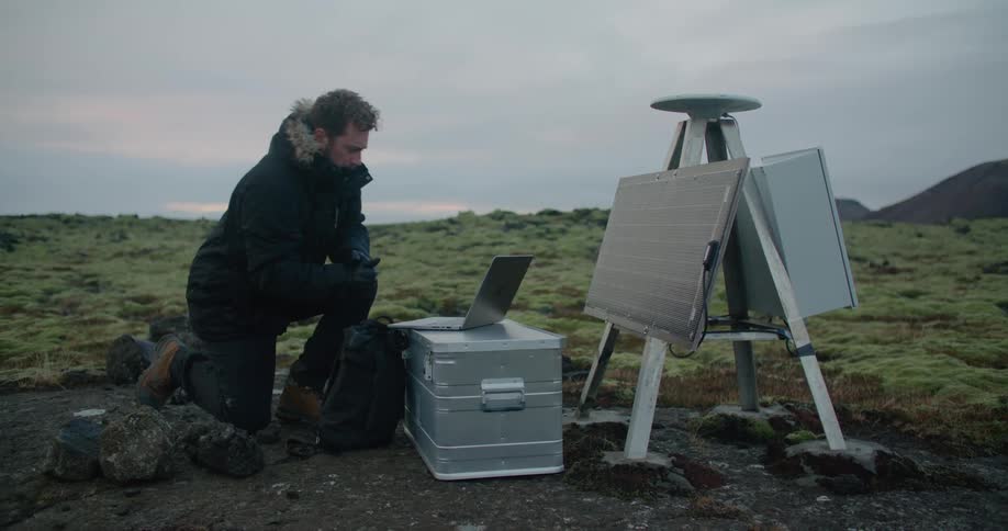A man is kneeling down in front of a laptop on a box near some scientific equipment