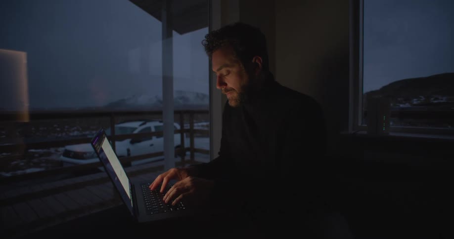 A man is sitting at a table in a dark room typing on a laptop computer with windows behind him