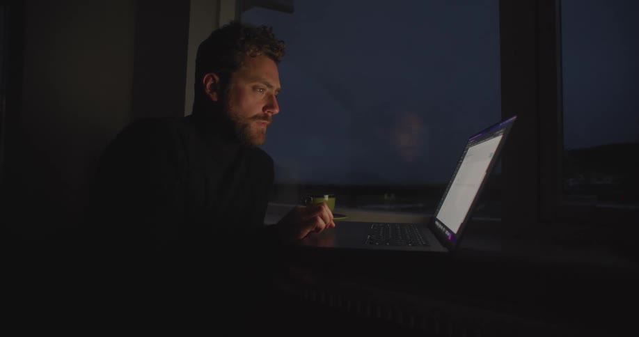 A man is sitting at a table in a dark room typing on a laptop computer