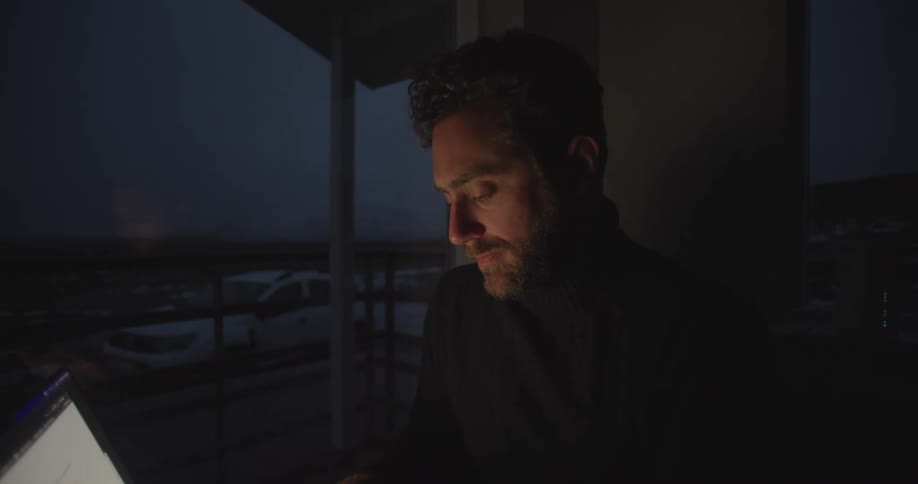 A man is sitting at a table in a dark room typing on a laptop computer