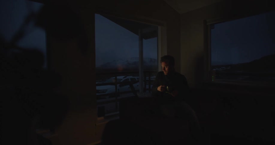 Man with beard and black turtleneck sweater drinks from cup and works on laptop in dark room