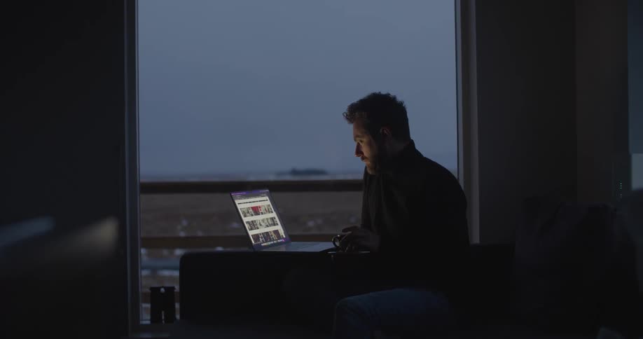 A man is sitting at a table near a window typing on a laptop computer