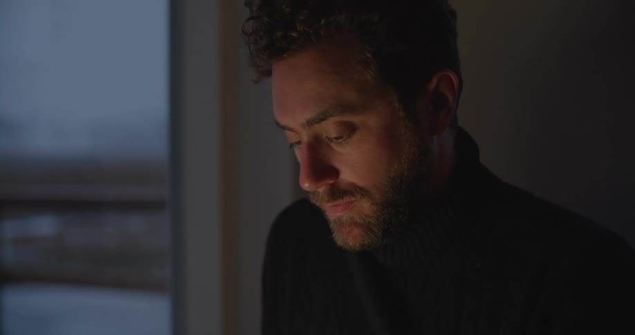Man with beard and black sweater works on laptop in dark room