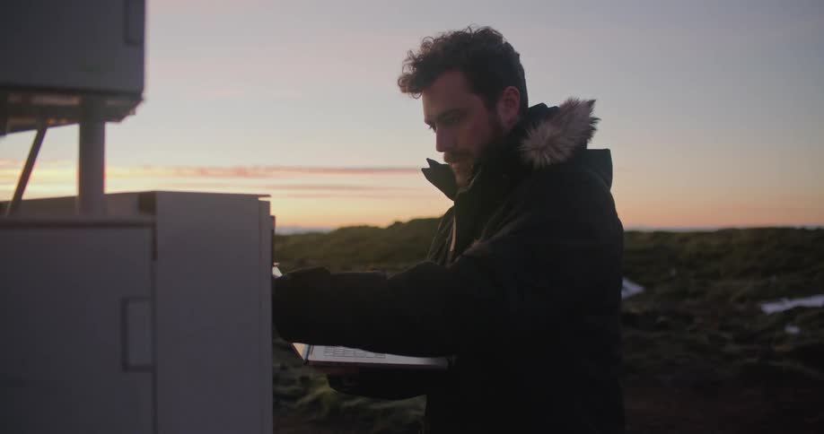 A man is standing outside in front of a utility box holding a laptop computer and typing with the other hand