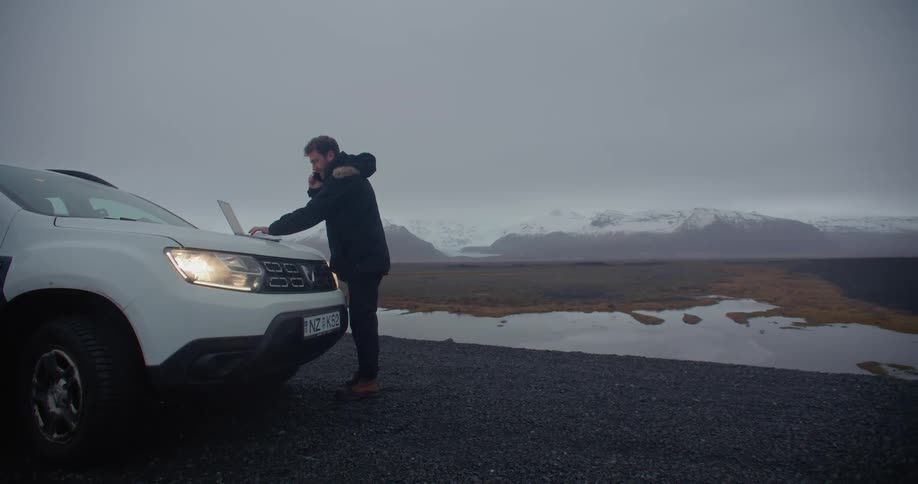 A man is standing outside wearing a winter coat and typing on a laptop computer that is sitting on the hood of a car as he talks on the phone