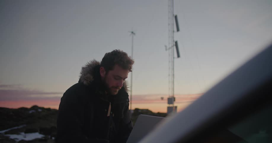 A man is standing outside wearing a winter coat and typing on a laptop computer that is sitting on the hood of a car with utility towers behind him