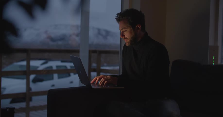 Man with beard and black turtleneck sweater works on laptop in dark room with white car and distant snowy mountain in the background