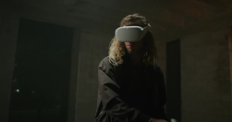 Blonde long haired man playing with a virtual reality game inside a room at night