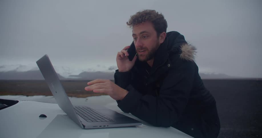 A man stands in a field with his laptop open on the hood of a car while he talks on a cell phone