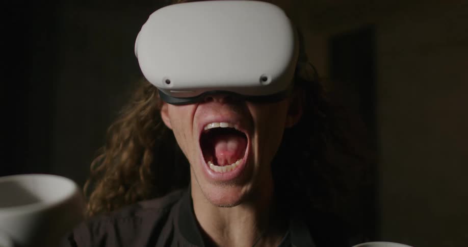 A man with long blond hair wearing a virtual reality headset screams as his hair is blown backwards