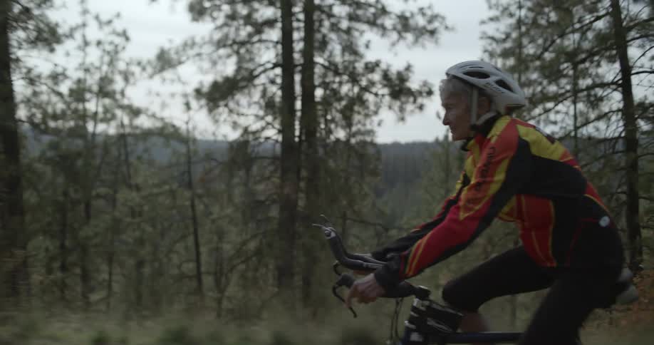 An elderly woman rides a bicycle along a path through a forested countryside during the day