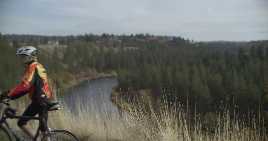 Cyclist wearing helmet walks bicycle along with river forest and town in the background