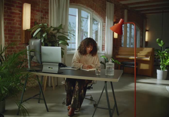 Woman sits at table writing while looking at laptop with piano in the background