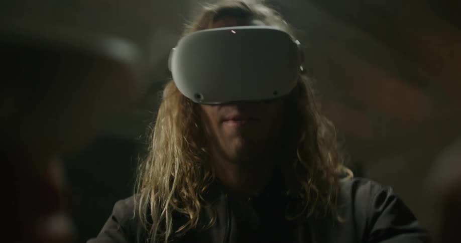 A man with long blond hair is wearing a virtual reality headset and playing a game