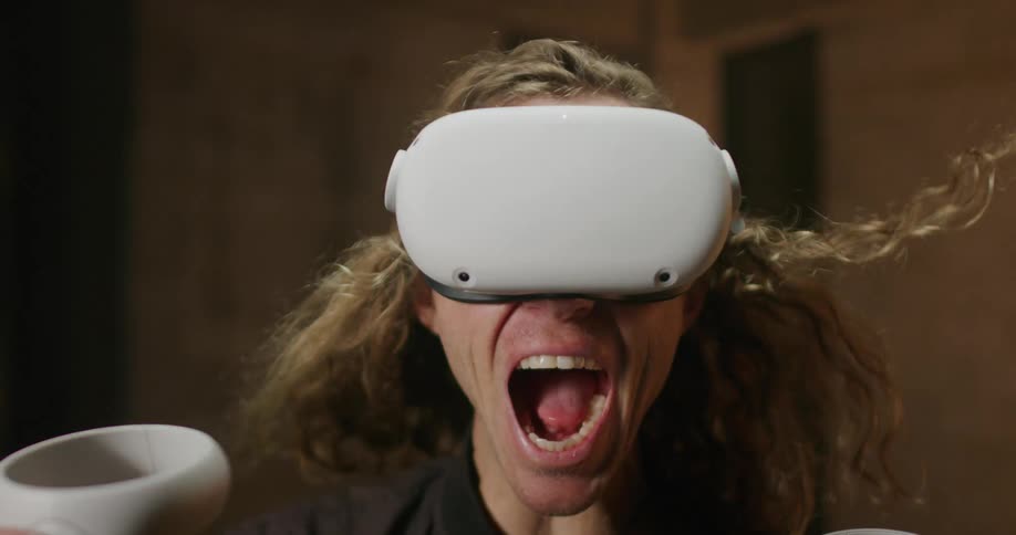 Blonde man wearing vr headset and holding game controllers yells as wind blows back his hair