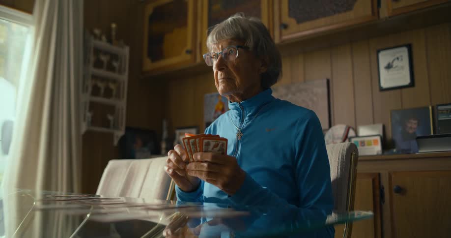 An elderly woman plays solitaire by herself at a table indoors during the day