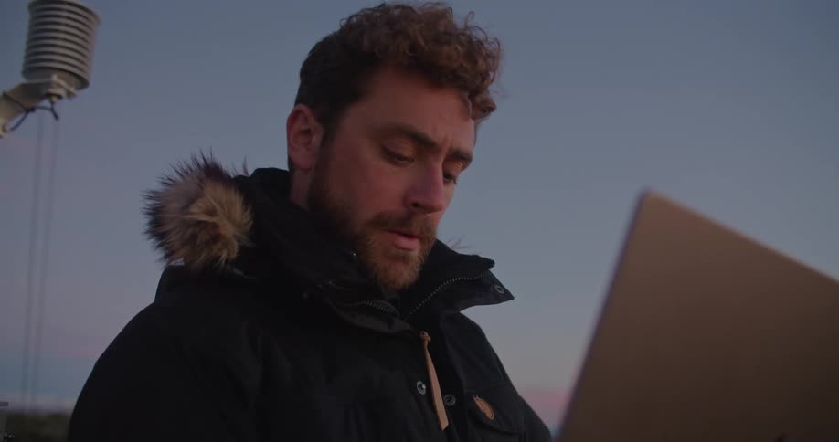 A man is standing outside wearing a winter coat typing on a laptop computer and there are utility towers behind him