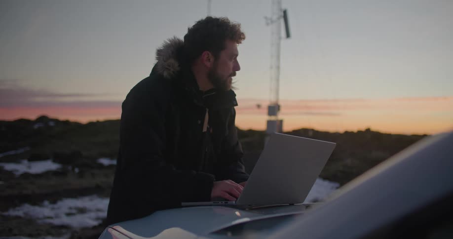 A man is standing outside wearing a winter coat and typing on a laptop computer that is sitting on the hood of a car with utility towers nearby