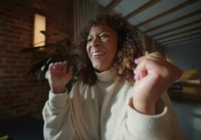 Woman with brown wavy hair and beige turtleneck sweater makes video call on laptop laughing and talking next to brick wall in room