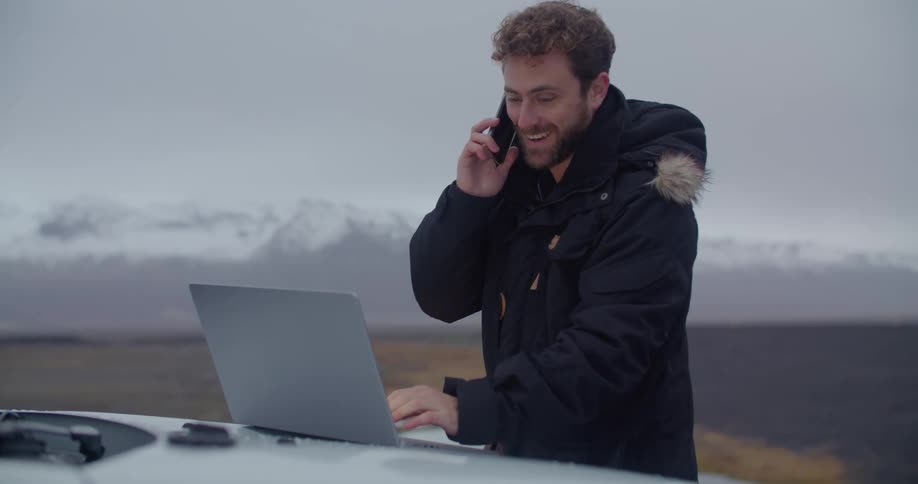 A man is standing outside wearing a winter coat and typing on a laptop computer that is sitting on the hood of a car as he talks on the phone
