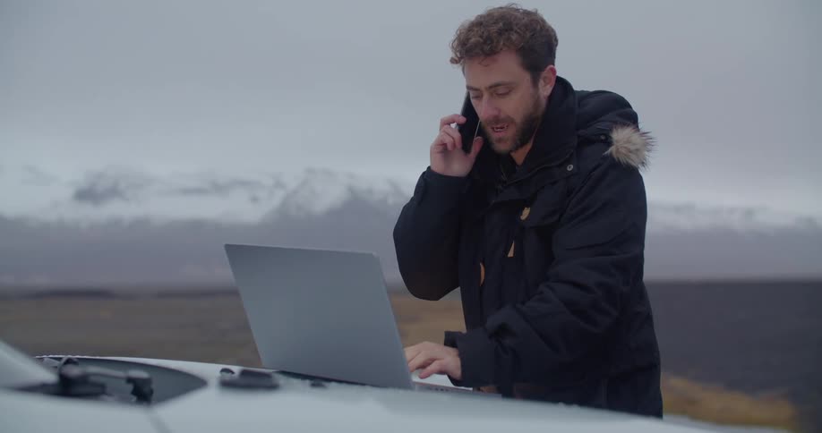 A man wearing a winter coat is typing on a laptop computer sitting on the hood of an automobile as he talks on the phone