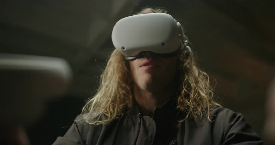 A man with long blond hair is wearing a virtual reality headset and playing a game