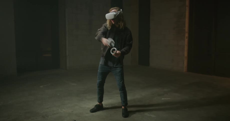 A man with long blond hair wearing virtual reality goggles is holding controllers and swinging his arms as he plays a game