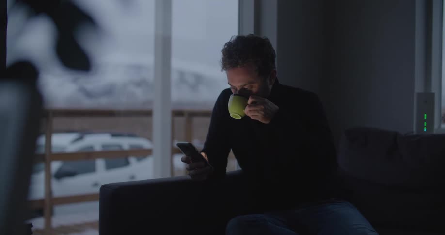 Man sitting near a window drinking coffee staring at a cell phone