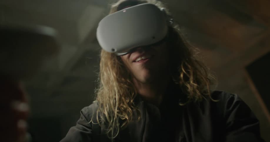 A man with long blond hair is wearing a virtual reality headset and playing a game