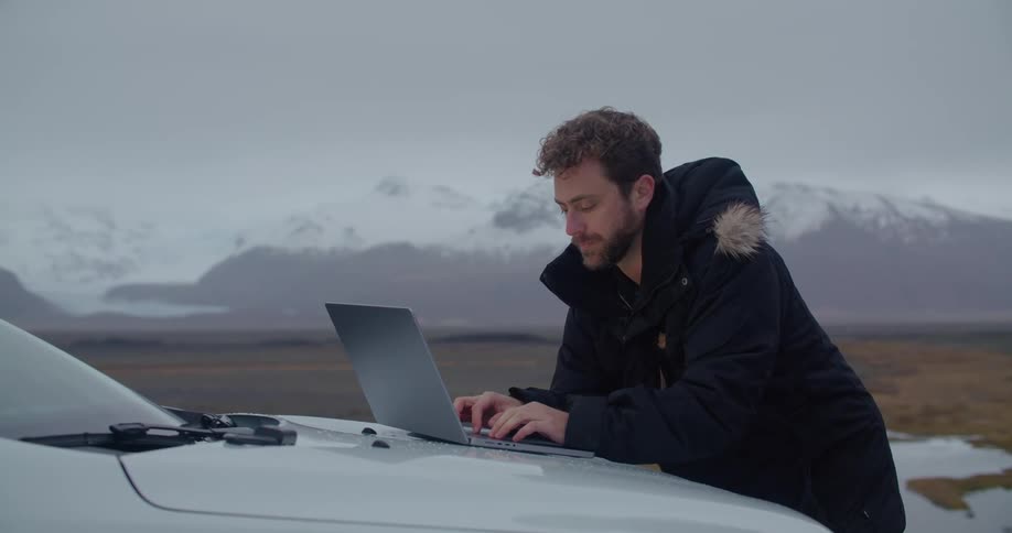 A man is standing outside wearing a winter coat and typing on a laptop computer that is sitting on the hood of a car