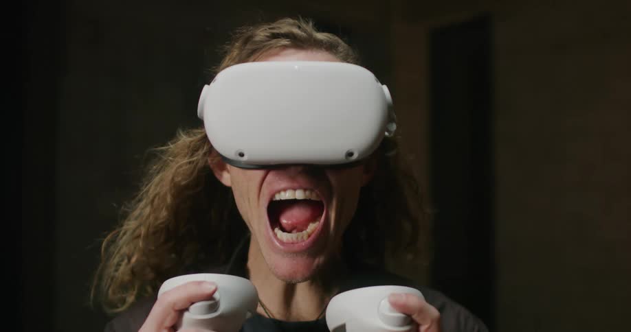 A man with long blond hair wearing a virtual reality headset and playing a game screams as his hair is blown backwards
