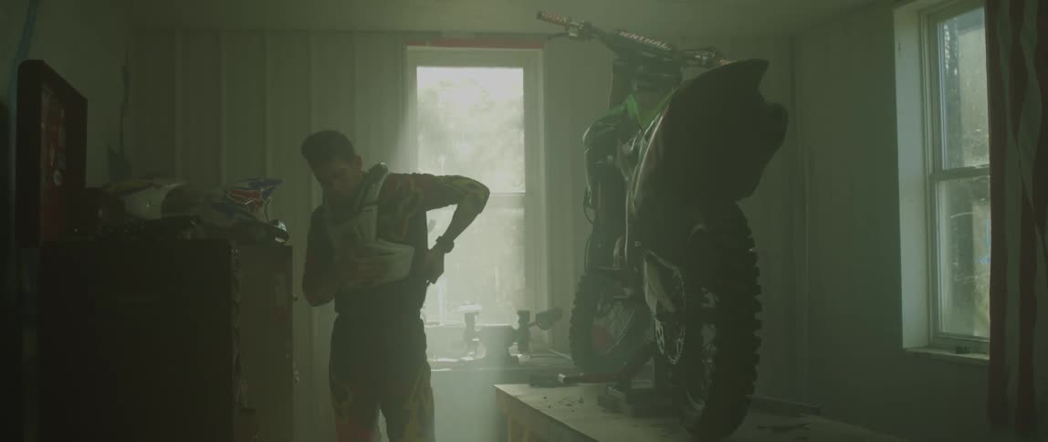 A racer puts on a protective vest while in a garage with his motorcycle on a workbench