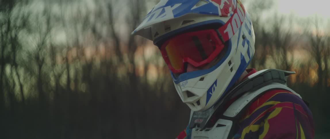 Man wearing a colorful motorcycle motocross helmet stands in front of a treeline at sunset