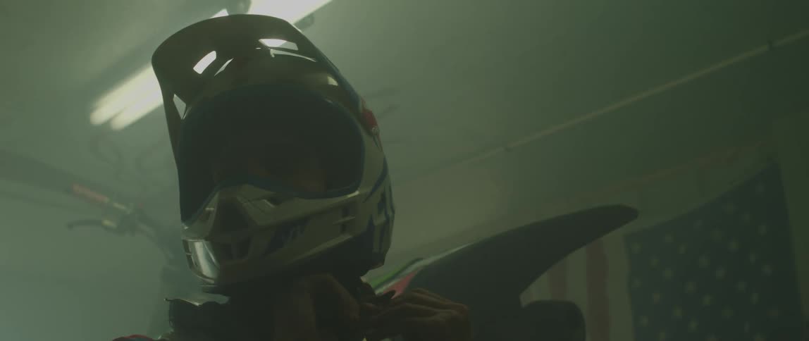 A man is indoors next to a motorcycleas he straps on a helmet
