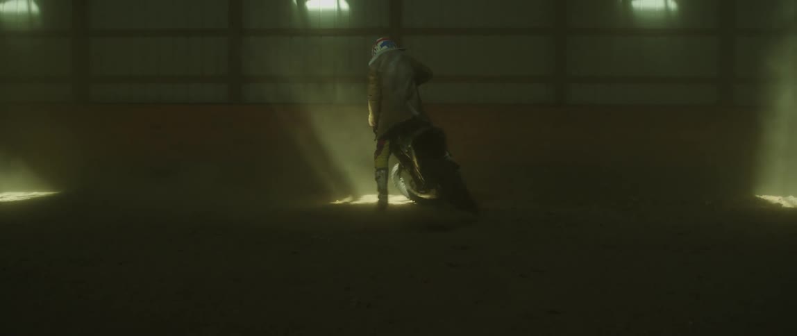 Motorcyclist rides bike in circle in large dim building lit by streaks of light from outside