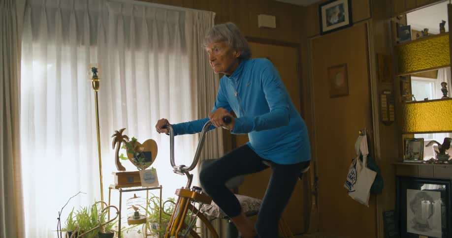 An elder woman pedals on an exercise bike in the living room of a house during the day